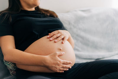 Unrecognizable pregnant woman hugging tummy at home sitting on sofa