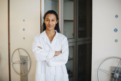 Portrait of confident young engineering student standing with arms crossed against storage cabinets in university