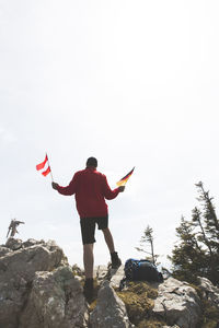 Rear view of mature man holding national flags while standing on mountain against sky