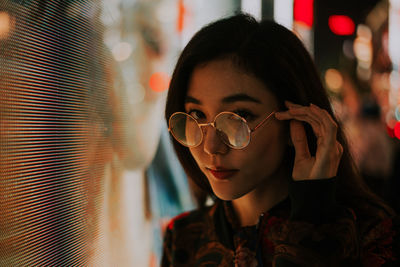 Close-up portrait of woman standing by illuminated lighting at night in city