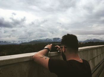 Rear view of man photographing mountains with mobile phone while standing by retaining wall against cloudy sky