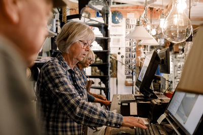 Confident senior female owner using computer while standing amidst coworkers at checkout in illuminated hardware store