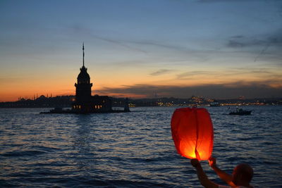 Illuminated building by sea against sky during sunset