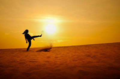 Silhouette woman kicking sand at beach against orange sky during sunset