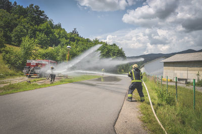 Firefighter spraying water on road against cloudy sky