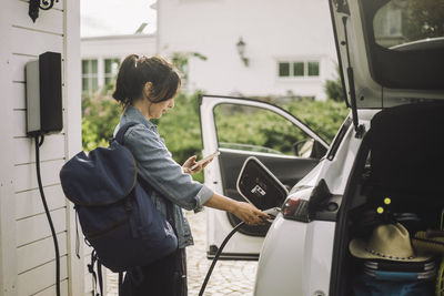 Side view of woman using smart phone while charging electric car