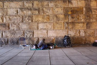 Homeless person sitting on sidewalk against stone wall