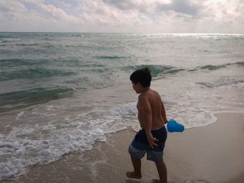 Full length of shirtless boy with bucket walking at beach