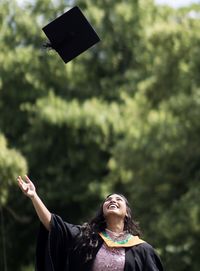 Woman throwing mortarboard in air against trees