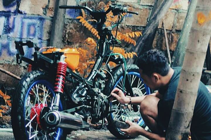 bicycle, mode of transportation, transportation, land vehicle, one person, motorcycle, repairing, occupation, wheel, pedal, mechanic, adult, men, casual clothing, technician, young adult, day, workshop, tire, outdoors, garage, bicycle shop, spoke