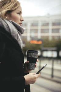 Side view of businesswoman holding mobile phone and disposable coffee cup at tram station