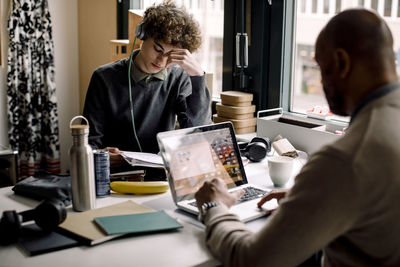 Boy wearing headphones studying while sitting with businessman using laptop at desk