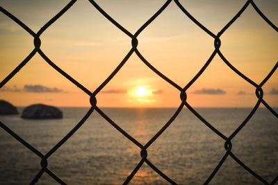 Close-up of chainlink fence in front of sea at sunset