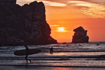 Side view of silhouette man carrying surfboard while walking at shore during sunset
