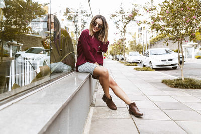 Portrait of smiling young woman sitting on sidewalk by glass building