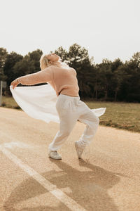 Blond female in casual clothes looking up while dancing with white sheet on asphalt road near green forest in sunlight