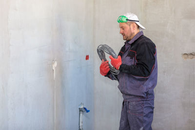 An electrician stands against the wall with an electrical cable for sockets.