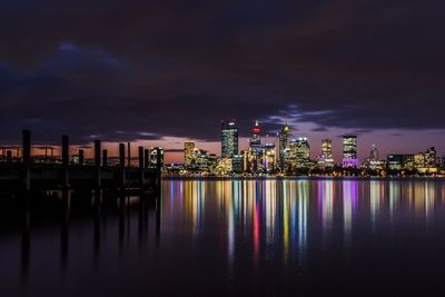 Illuminated buildings by river against sky at night