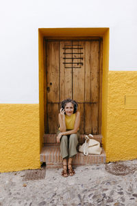 Mature woman listening music through headphone while sitting against old door