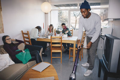 Man cleaning floor while friends sitting in college dorm room