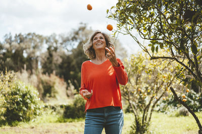 Happy mature woman juggling oranges standing by tree in garden