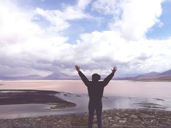 Rear view of man with arms raised standing by laguna colorada against cloudy sky