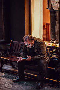 Full length of man using mobile phone while sitting on bench outside store