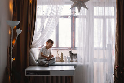 Cute boy playing with kitten sitting on the windowsill near the window. cozy home.