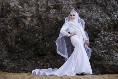 Portrait of beautiful bride standing against rock formation at beach