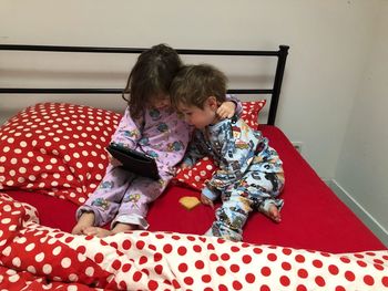 Siblings using digital tablet while sitting on bed at home