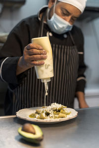 Crop middle aged ethnic male cook pouring white sauce from bottle on delicious pasta with grated cheese in restaurant kitchen