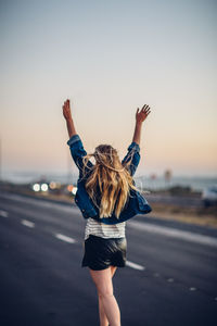 Rear view of young woman with arms raised walking on road against clear sky during sunset