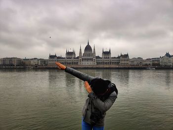 Man gesturing by river against building and sky