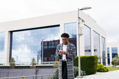From below of happy young african american male student in stylish outfit browsing smartphone while standing on city street near modern building