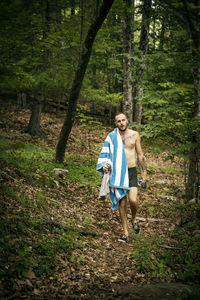 Shirtless man carrying towel and clothes while walking in forest
