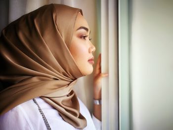 Side view of young woman in hijab looking through window at home