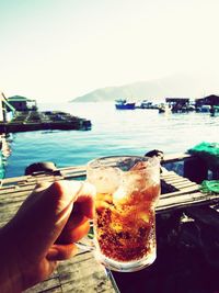 Cropped hand holding beer by lake against clear sky