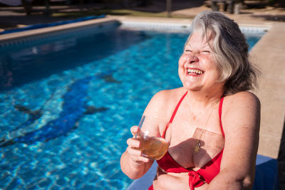 High angle of delighted senior female tourist in bikini laughing brightly while chilling on poolside with drink