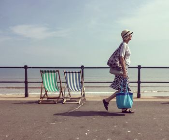 Side view of woman walking with bags on promenade by sea against sky