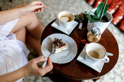A girl in a white robe eats tiramisu, standing on the table next to two cups of coffee