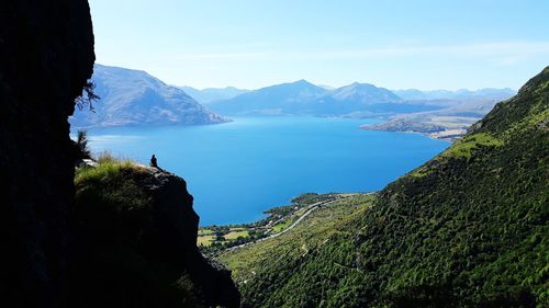 Mid distant view of woman sitting on cliff against lake and sky