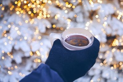 Cropped hand of person holding coffee cup against illuminated lights during winter