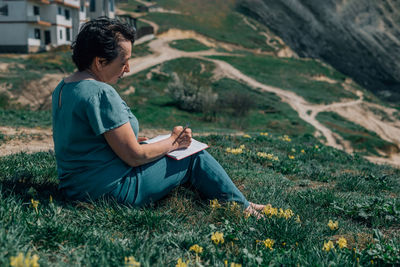 Adult woman thoughtfully writes plans diary on a sunny day on a mountain against backdrop of the sea