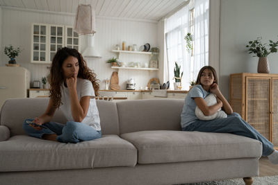 Upset quarreled woman and girl sit on opposite sides of sofa in living room look back at each other