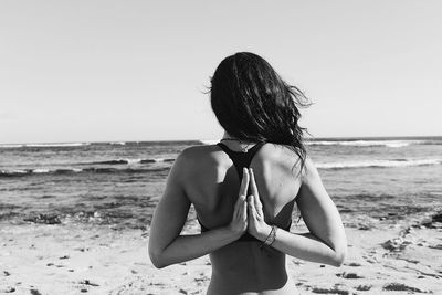 Rear view of woman with hands behind back meditating at beach against sky