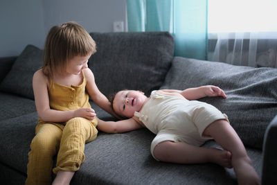 Cute european children in overalls play together relaxing on gray sofa in bright minimalist 