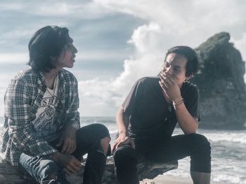 Young men sitting on rock at beach against sky