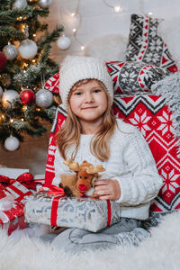 Happy little girl in white hat holding christmas gift boxes while sitting on floor.