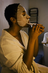 Young woman removing facial mask on face while sitting in bathroom
