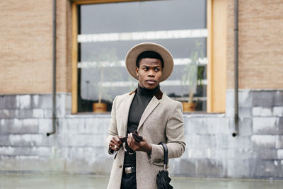 Young man holding hat standing against built structure